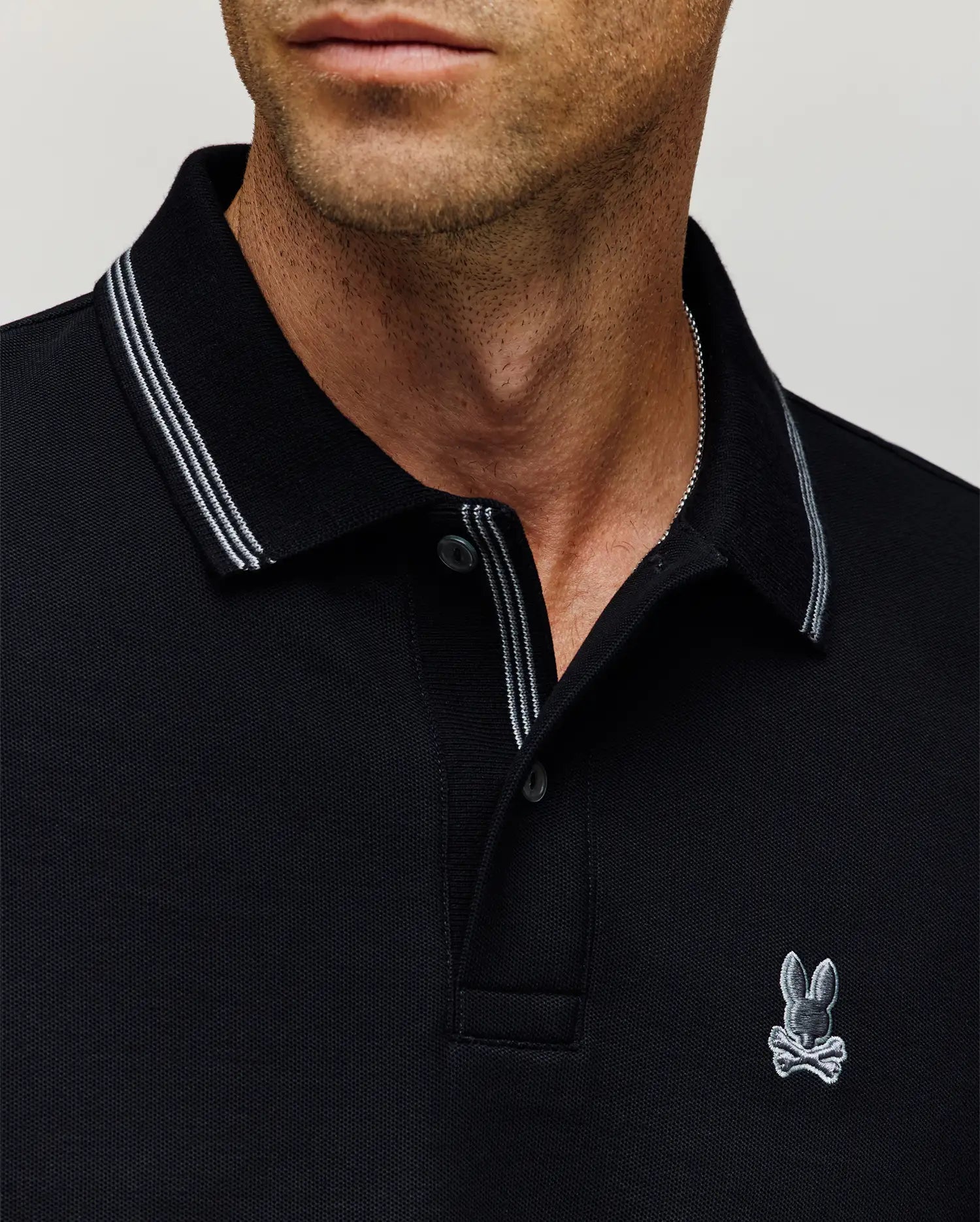 A man wears the Psycho Bunny MENS HART PIQUE POLO SHIRT (MT0100032), a black designer polo with white trim and a small bunny with crossbones logo on the chest; image shows only his lower face, neck, and upper torso. (2)