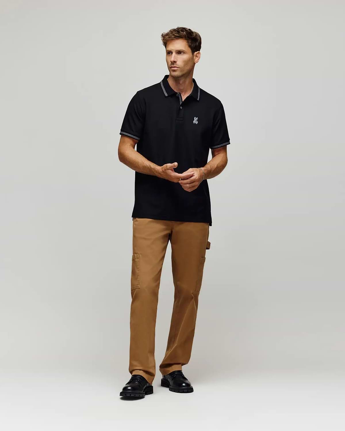 A man stands against a plain background wearing the Psycho Bunny MENS HART PIQUE POLO SHIRT (MT0100032), tan cargo pants, and black shoes. He faces forward, hands relaxed in front of him. (4)