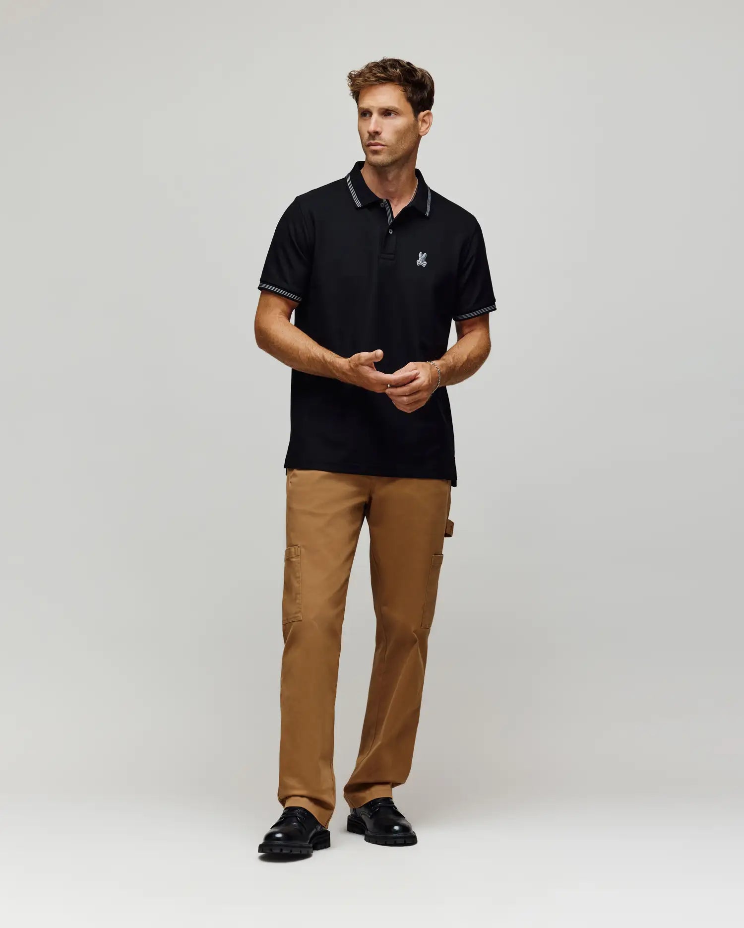 A man stands against a plain background wearing the Psycho Bunny MENS HART PIQUE POLO SHIRT (MT0100032), tan cargo pants, and black shoes. He faces forward, hands relaxed in front of him. (4)