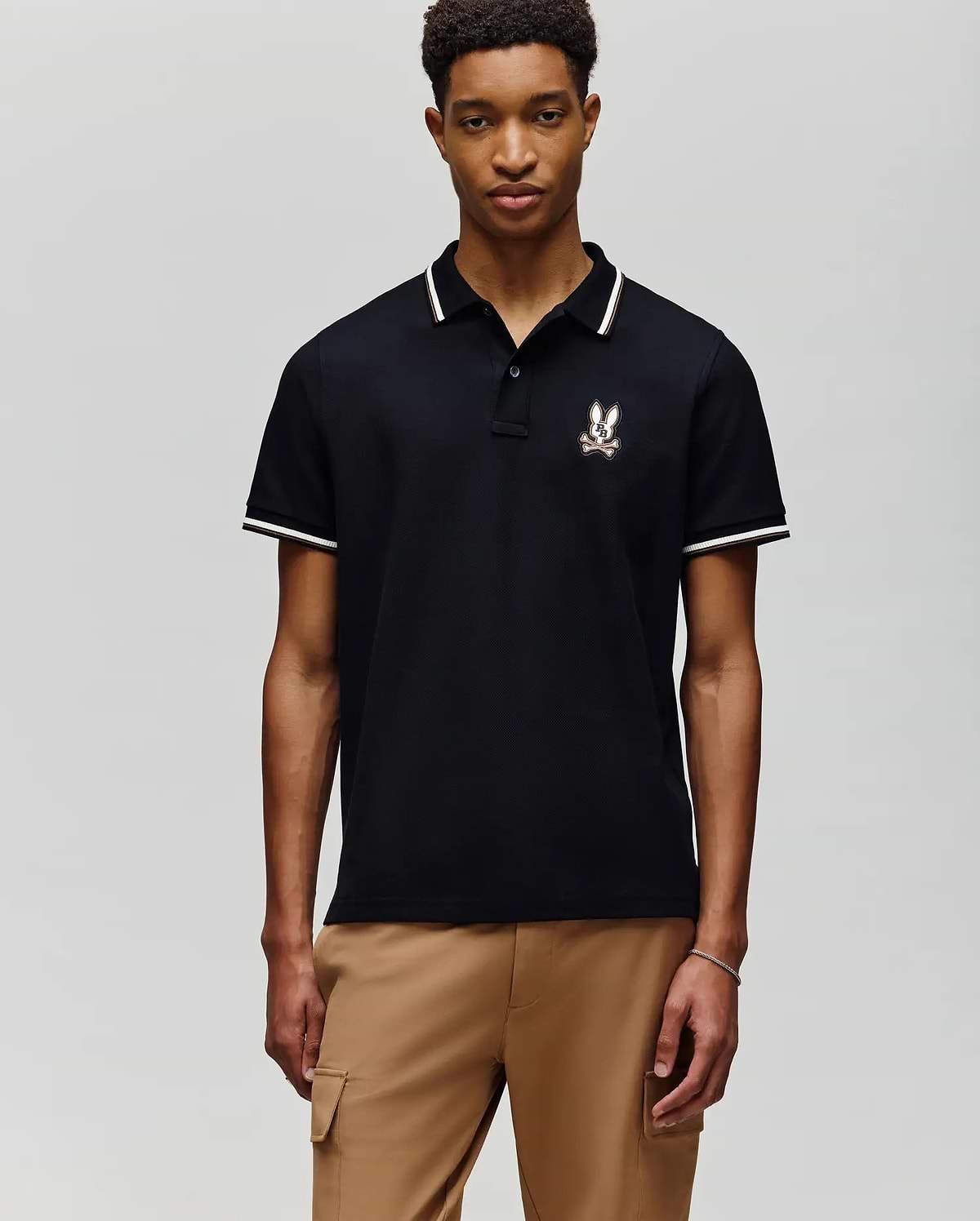 A young man faces forward against a gray background, wearing Psycho Bunny's MENS WADE PIQUE POLO - MT0100044 in black with white trim and an embroidered crest on the chest, paired with light brown pants.