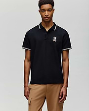 A young man faces forward against a gray background, wearing Psycho Bunny's MENS WADE PIQUE POLO - MT0100044 in black with white trim and an embroidered crest on the chest, paired with light brown pants.