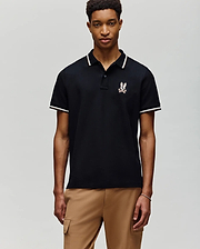 A young man faces forward against a gray background, wearing Psycho Bunny's MENS WADE PIQUE POLO - MT0100044 in black with white trim and an embroidered crest on the chest, paired with light brown pants.
