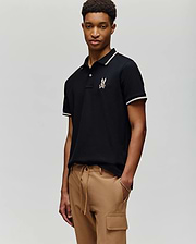 A young man stands against a plain background wearing the Psycho Bunny MENS WADE PIQUE POLO - MT0100044 in black with white trim and a chest logo, paired with tan cargo pants.