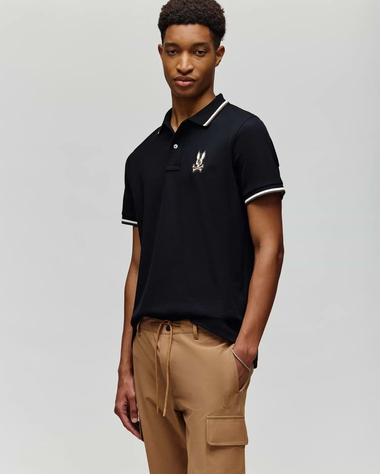 A young man stands against a plain background wearing the Psycho Bunny MENS WADE PIQUE POLO - MT0100044 in black with white trim and a chest logo, paired with tan cargo pants.