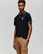 A young man stands against a plain background wearing the Psycho Bunny MENS WADE PIQUE POLO - MT0100044 in black with white trim and a chest logo, paired with tan cargo pants.