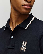 A person wears the Psycho Bunny MENS WADE PIQUE POLO (MT0100044), a dark men's shirt with a striped collar and the brand's signature cartoon bunny head above crossed bones patch on the chest. The face is partially visible against a plain background.