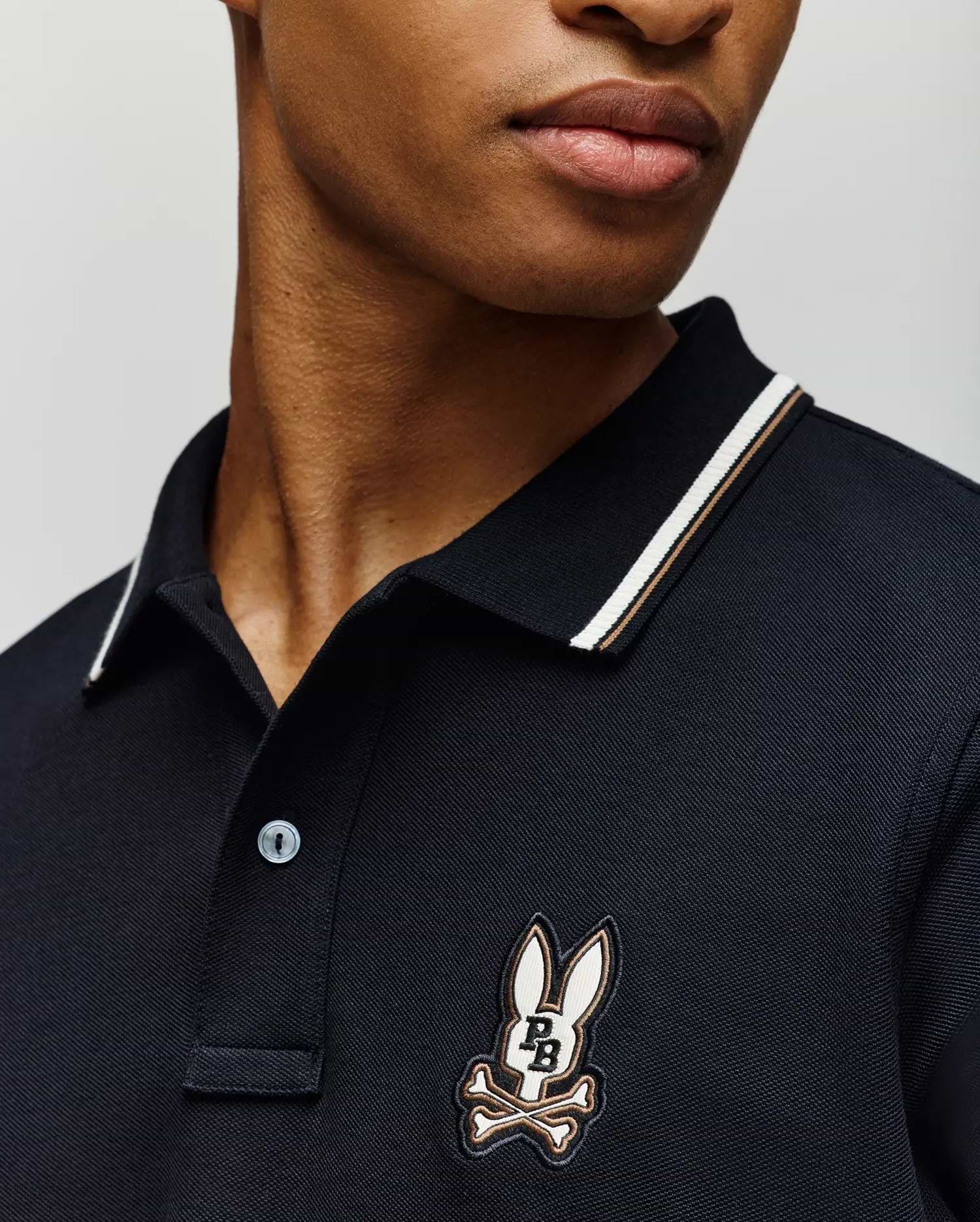 A person wears the Psycho Bunny MENS WADE PIQUE POLO (MT0100044), a dark men's shirt with a striped collar and the brand's signature cartoon bunny head above crossed bones patch on the chest. The face is partially visible against a plain background.