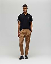 A person stands against a plain background wearing the Psycho Bunny MENS WADE PIQUE POLO - MT0100044 in classic black with a white logo, paired with tan drawstring pants and black sneakers featuring white stripes.