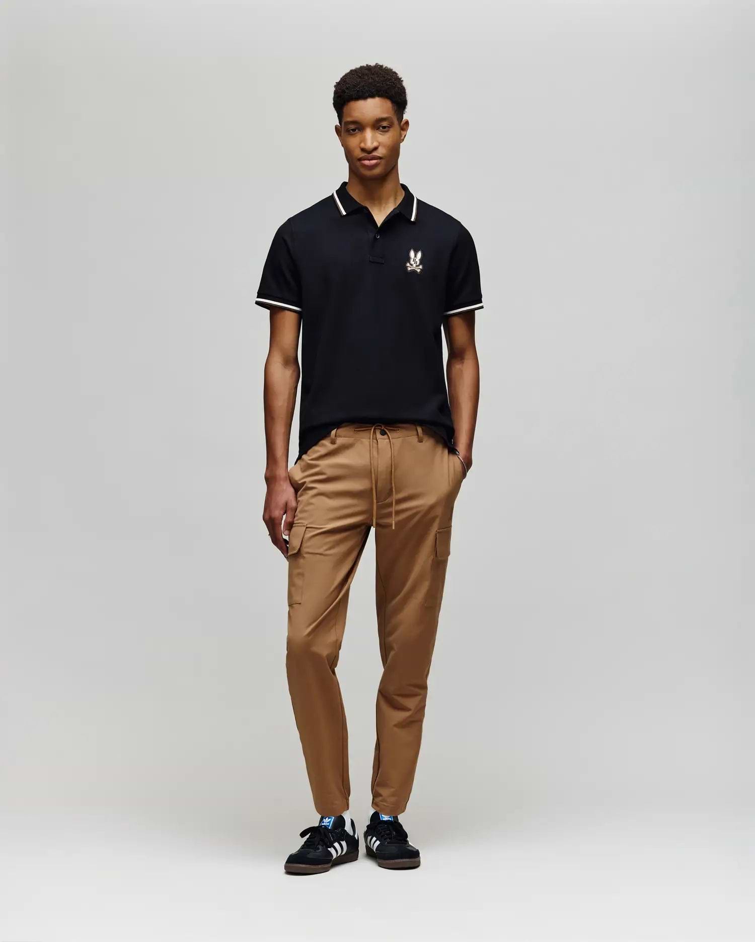 A person stands against a plain background wearing the Psycho Bunny MENS WADE PIQUE POLO - MT0100044 in classic black with a white logo, paired with tan drawstring pants and black sneakers featuring white stripes.