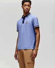 A young man stands against a light grey background wearing the MENS WADE PIQUE POLO (MT0100044) by Psycho Bunny in light blue, featuring an embroidered bunny logo on the chest, paired with tan cargo pants. (1)