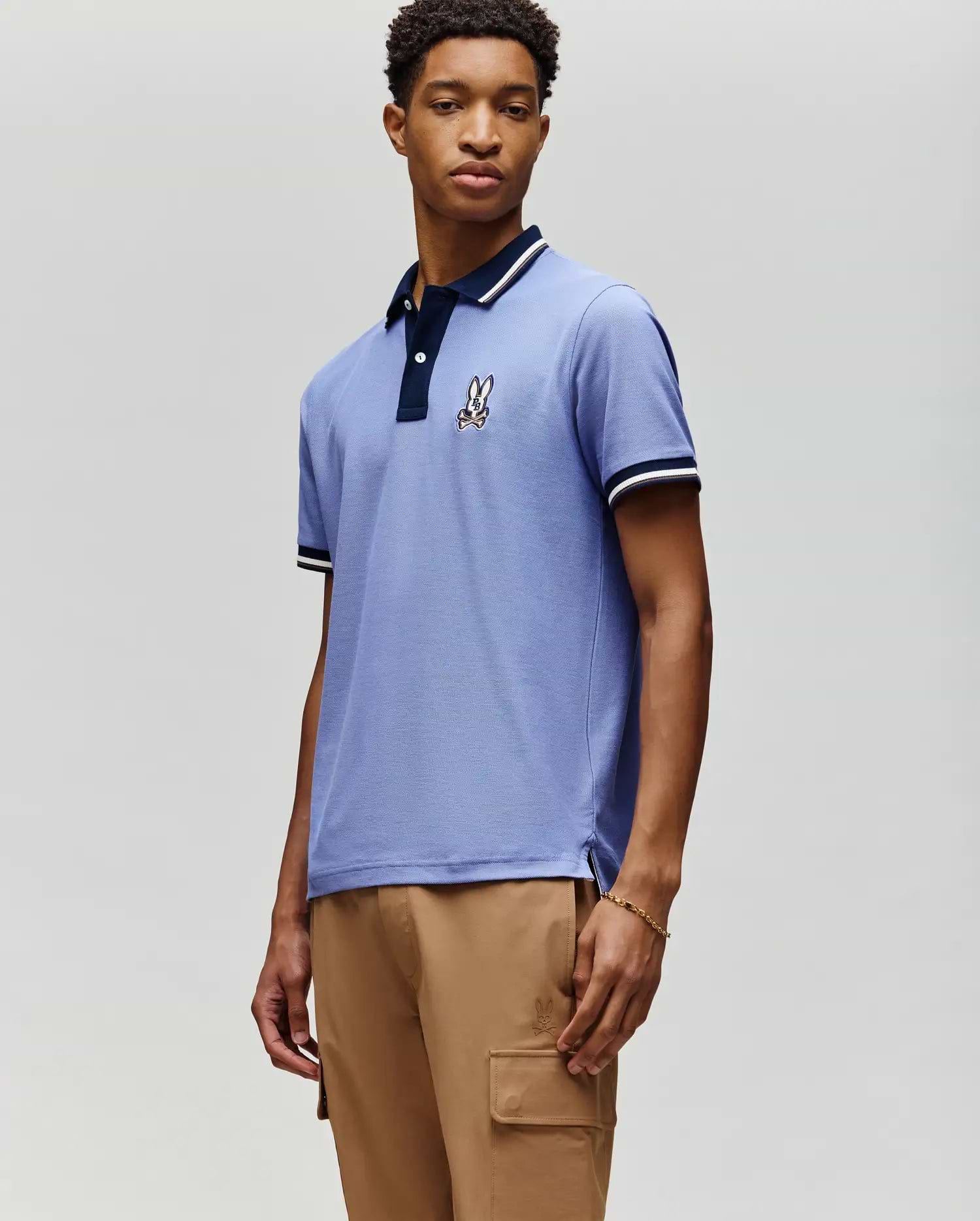 A young man stands against a light grey background wearing the MENS WADE PIQUE POLO (MT0100044) by Psycho Bunny in light blue, featuring an embroidered bunny logo on the chest, paired with tan cargo pants. (1)