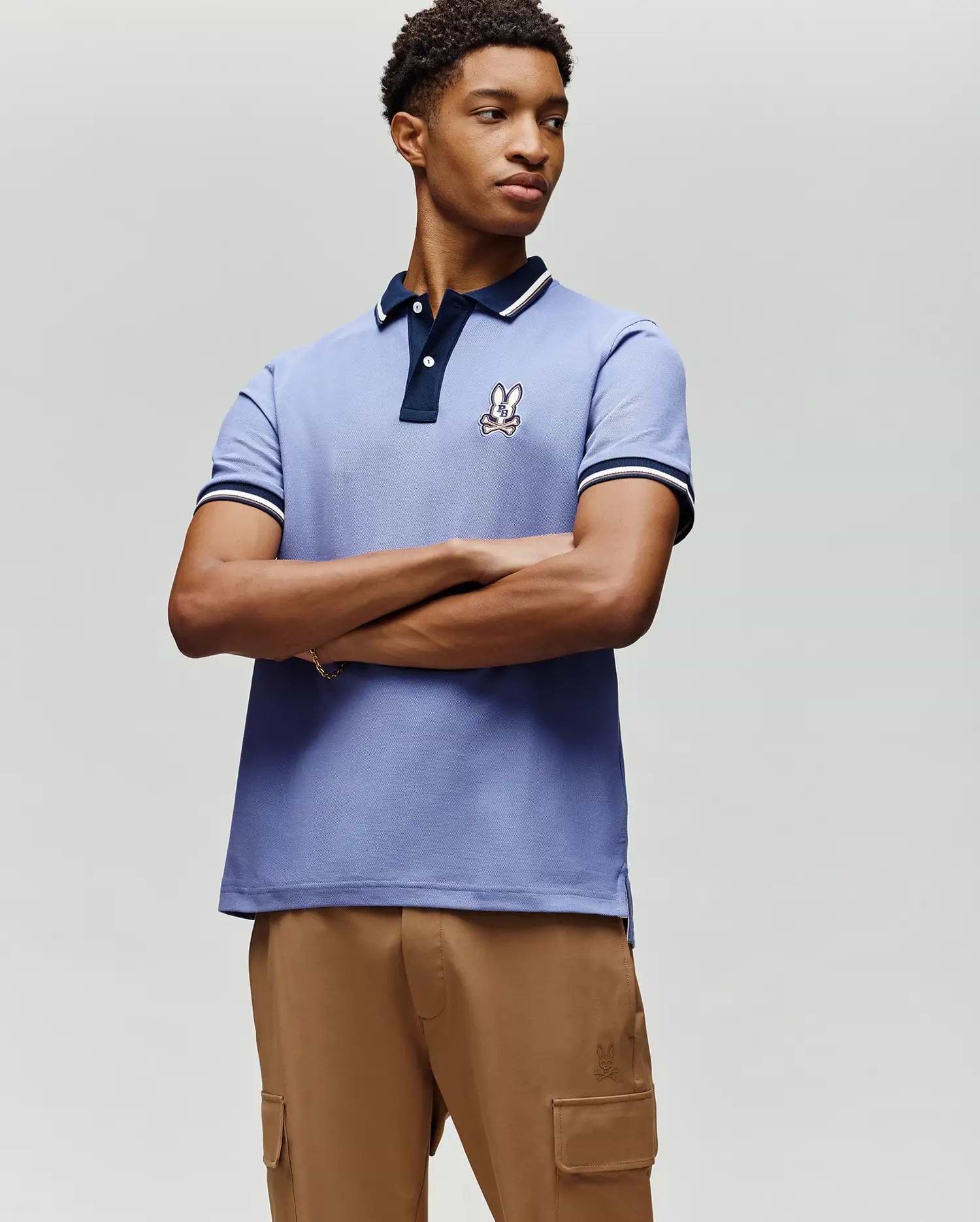 A young man stands with arms crossed in front of a plain light background, wearing the Psycho Bunny MENS WADE PIQUE POLO (MT0100044) in light blue with an embroidered bunny logo on the chest and tan cargo pants. (2)