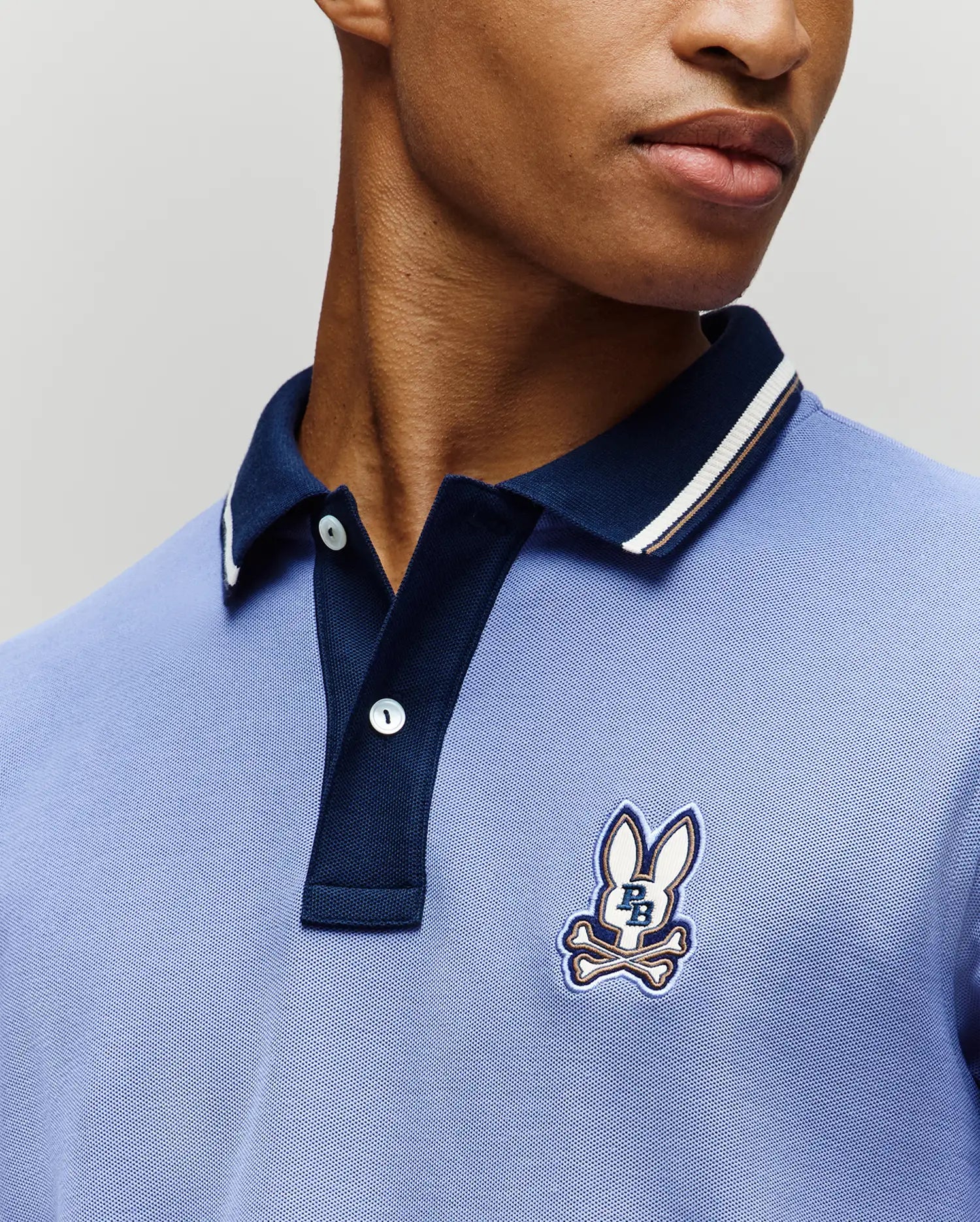 A person wears the Psycho Bunny MENS WADE PIQUE POLO (MT0100044), a light blue Pima cotton polo with a dark blue collar, colorblock tipping, and an embroidered bunny and crossbones logo. Only the lower face and upper torso are visible. (3)