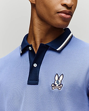 A person wears the Psycho Bunny MENS WADE PIQUE POLO (MT0100044), a light blue Pima cotton polo with a dark blue collar, colorblock tipping, and an embroidered bunny and crossbones logo. Only the lower face and upper torso are visible. (3)