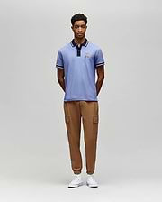 A man stands against a plain gray background, wearing the Psycho Bunny MENS WADE PIQUE POLO - MT0100044 in light blue, paired with tan cargo jogger pants and white sneakers. His hands are behind his back as he looks straight ahead. (4)