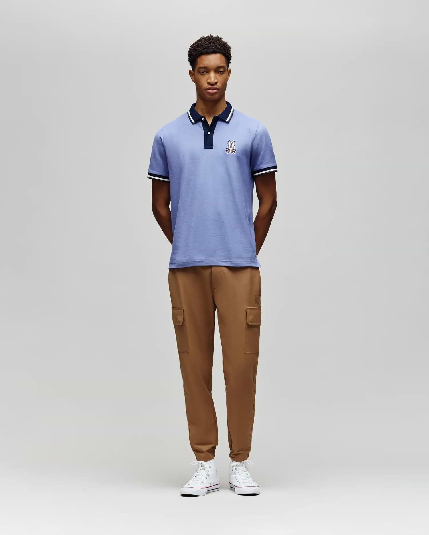 A man stands against a plain gray background, wearing the Psycho Bunny MENS WADE PIQUE POLO - MT0100044 in light blue, paired with tan cargo jogger pants and white sneakers. His hands are behind his back as he looks straight ahead. (4)