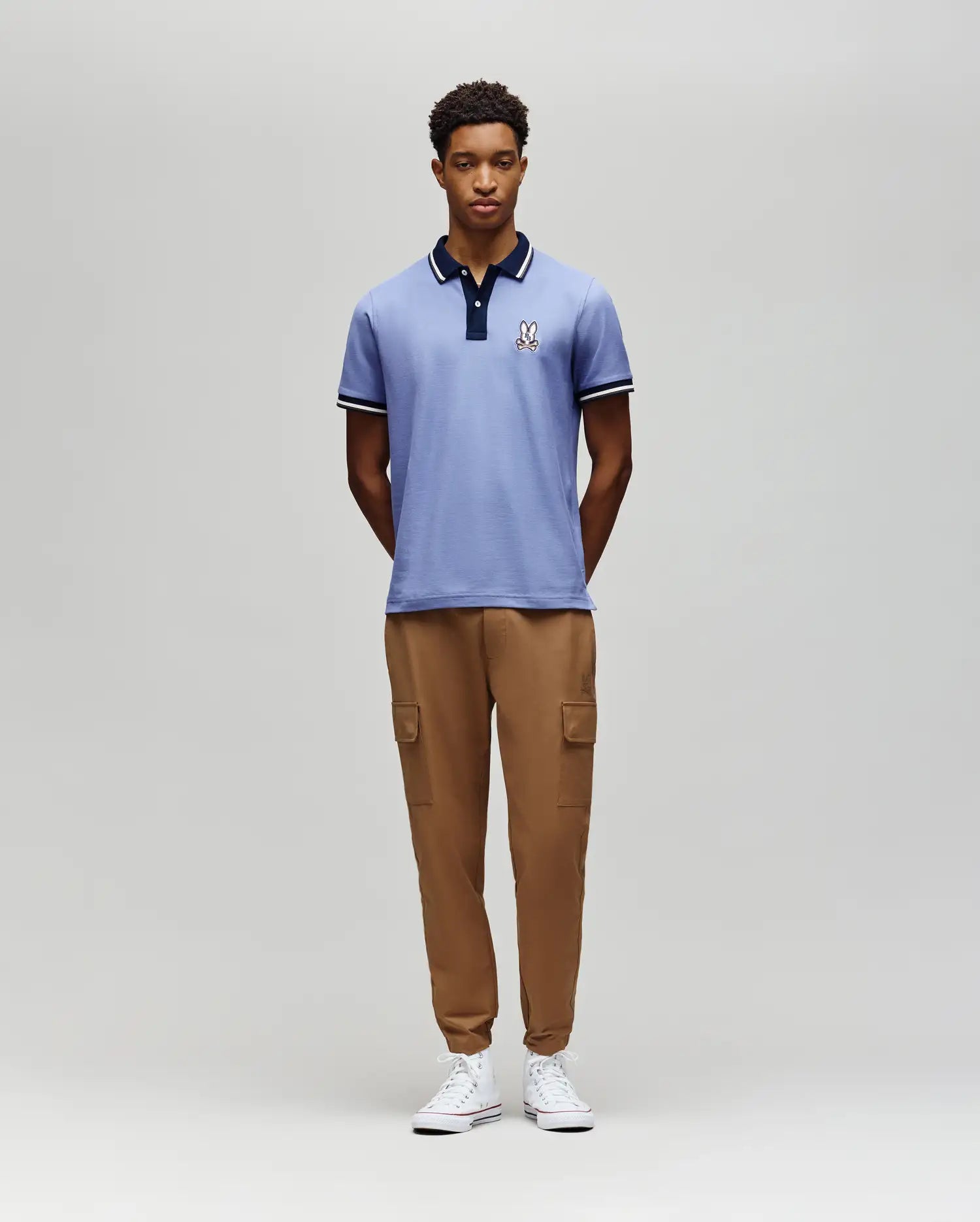 A man stands against a plain gray background, wearing the Psycho Bunny MENS WADE PIQUE POLO - MT0100044 in light blue, paired with tan cargo jogger pants and white sneakers. His hands are behind his back as he looks straight ahead. (4)