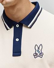 A close-up shows a person wearing the Psycho Bunny MENS WADE PIQUE POLO (MT0100044), a colorblock style with a navy collar, buttons, and an embroidered bunny logo above crossed bones plus "P B" on the left chest.