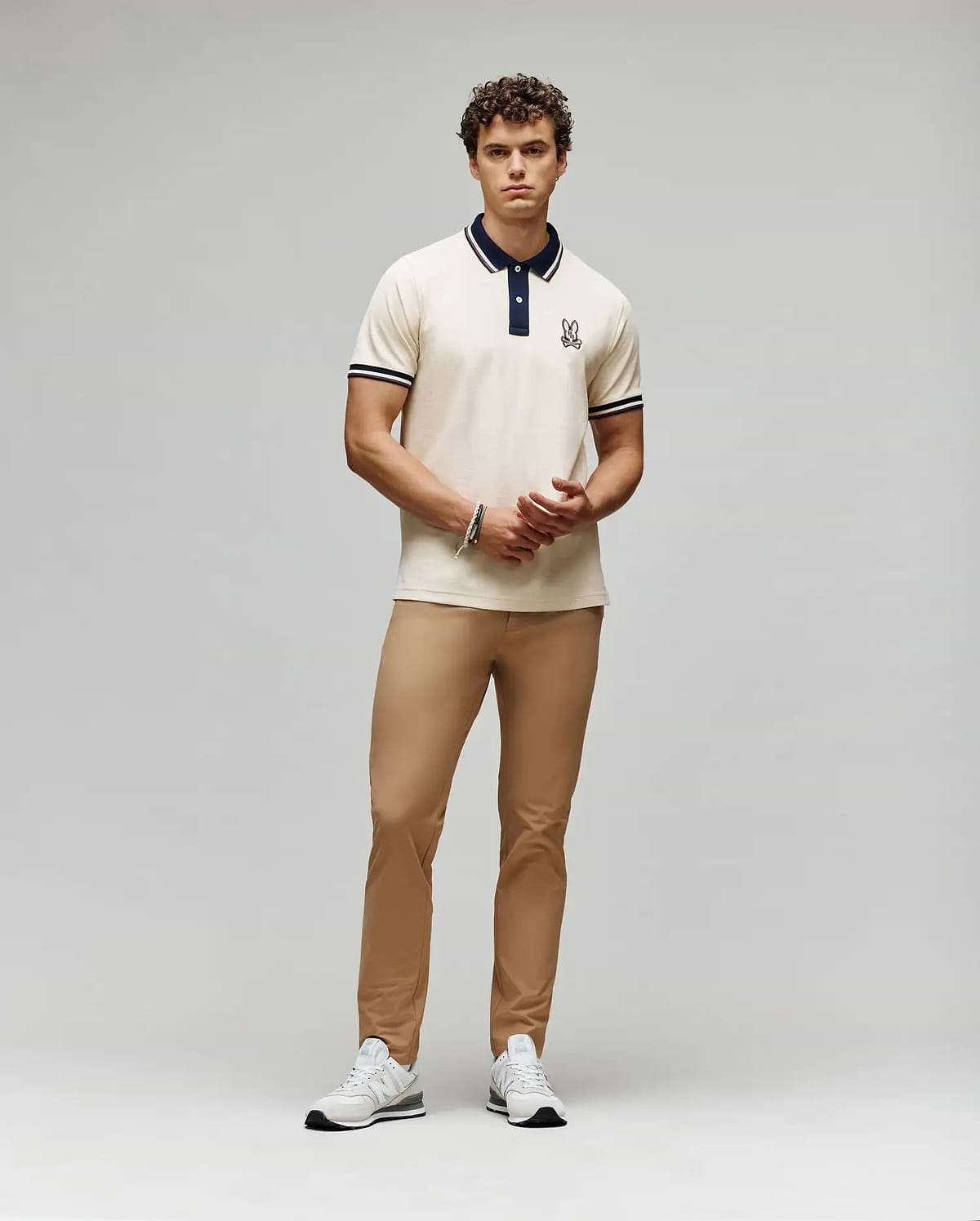 A man with curly hair and a neutral expression stands against a plain background wearing the Psycho Bunny MENS WADE PIQUE POLO - MT0100044, tan pants, and white sneakers. His hands are loosely clasped.