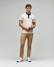 A man with curly hair and a neutral expression stands against a plain background wearing the Psycho Bunny MENS WADE PIQUE POLO - MT0100044, tan pants, and white sneakers. His hands are loosely clasped.