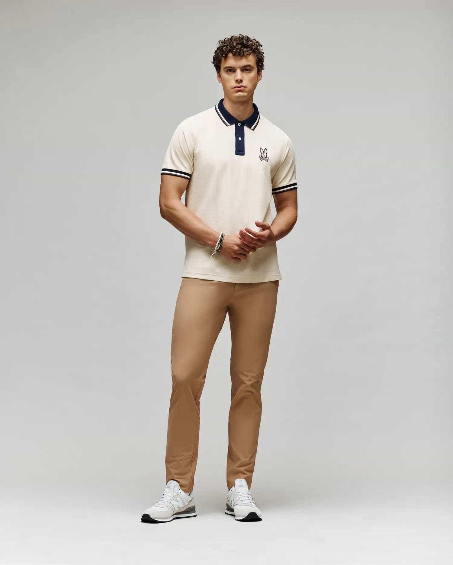 A man with curly hair and a neutral expression stands against a plain background wearing the Psycho Bunny MENS WADE PIQUE POLO - MT0100044, tan pants, and white sneakers. His hands are loosely clasped.