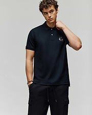 A young man with curly hair wears the Psycho Bunny MENS AUGUSTE PIQUE POLO SHIRT - MT0100051 and black cargo pants, standing against a plain background, looking at the camera neutrally with his right hand touching his neck. (1)