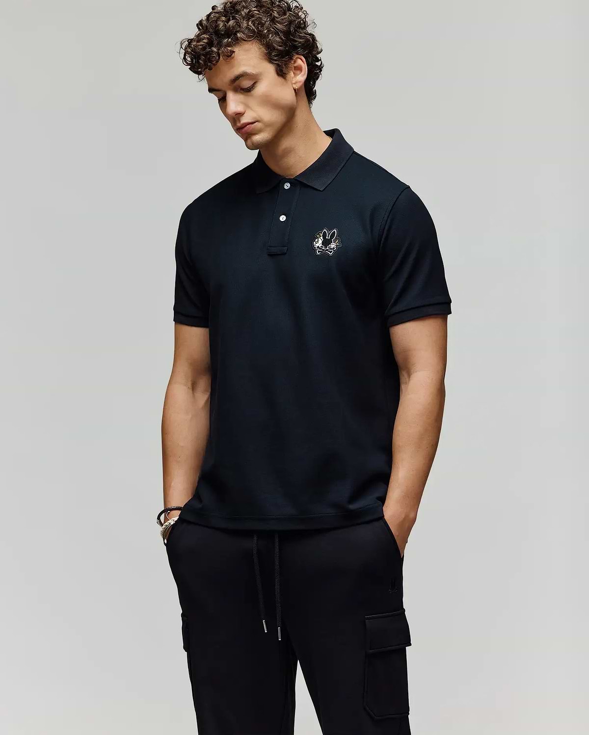 A man with curly hair wears the Psycho Bunny MENS AUGUSTE PIQUE POLO SHIRT - MT0100051 with an embroidered logo, styled varsity-like, and pairs it with black cargo pants. He stands hands-in-pockets, looking down against a plain background. (3)