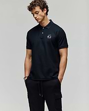 A man with curly hair wears the Psycho Bunny MENS AUGUSTE PIQUE POLO SHIRT - MT0100051 with an embroidered logo, styled varsity-like, and pairs it with black cargo pants. He stands hands-in-pockets, looking down against a plain background. (3)