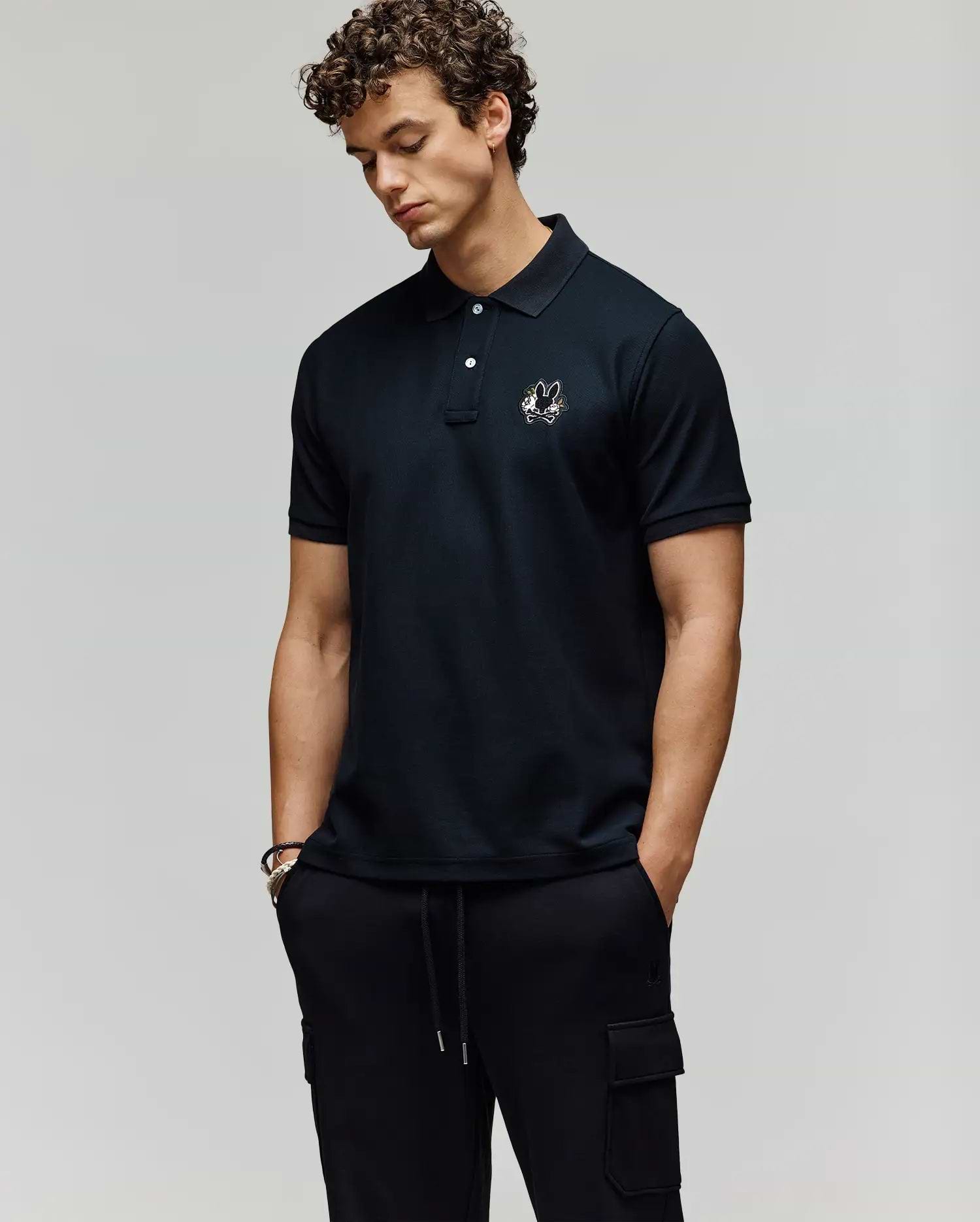 A man with curly hair wears the Psycho Bunny MENS AUGUSTE PIQUE POLO SHIRT - MT0100051 with an embroidered logo, styled varsity-like, and pairs it with black cargo pants. He stands hands-in-pockets, looking down against a plain background. (3)