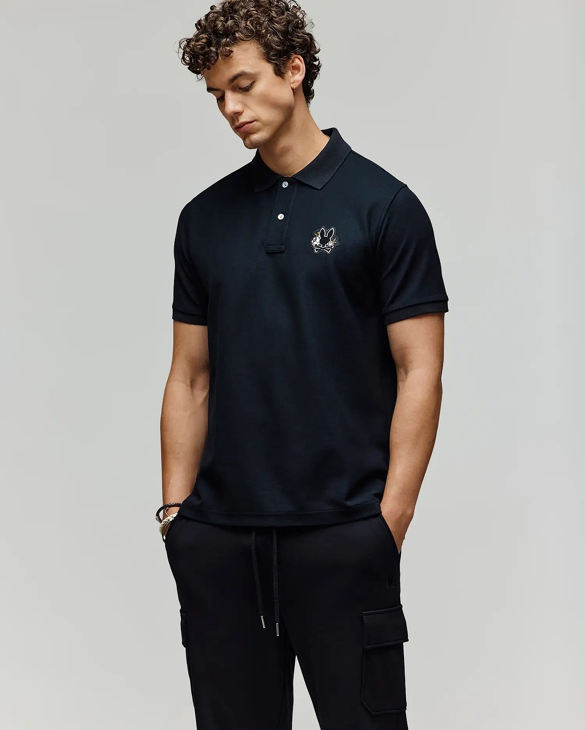 A man with curly hair wears the Psycho Bunny MENS AUGUSTE PIQUE POLO SHIRT - MT0100051 with an embroidered logo, styled varsity-like, and pairs it with black cargo pants. He stands hands-in-pockets, looking down against a plain background. (3)