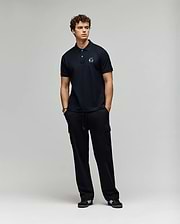 A young man with curly hair stands against a plain background, wearing a Psycho Bunny MENS AUGUSTE PIQUE POLO SHIRT - MT0100051 and black cargo pants. Black sneakers with white accents complete his varsity-inspired look as he gazes to the side. (4)
