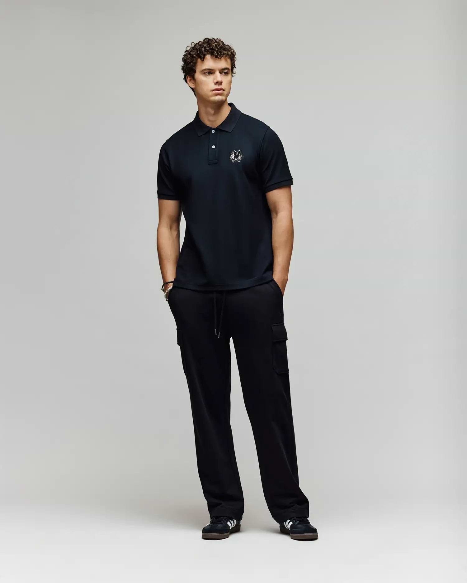 A young man with curly hair stands against a plain background, wearing a Psycho Bunny MENS AUGUSTE PIQUE POLO SHIRT - MT0100051 and black cargo pants. Black sneakers with white accents complete his varsity-inspired look as he gazes to the side. (4)