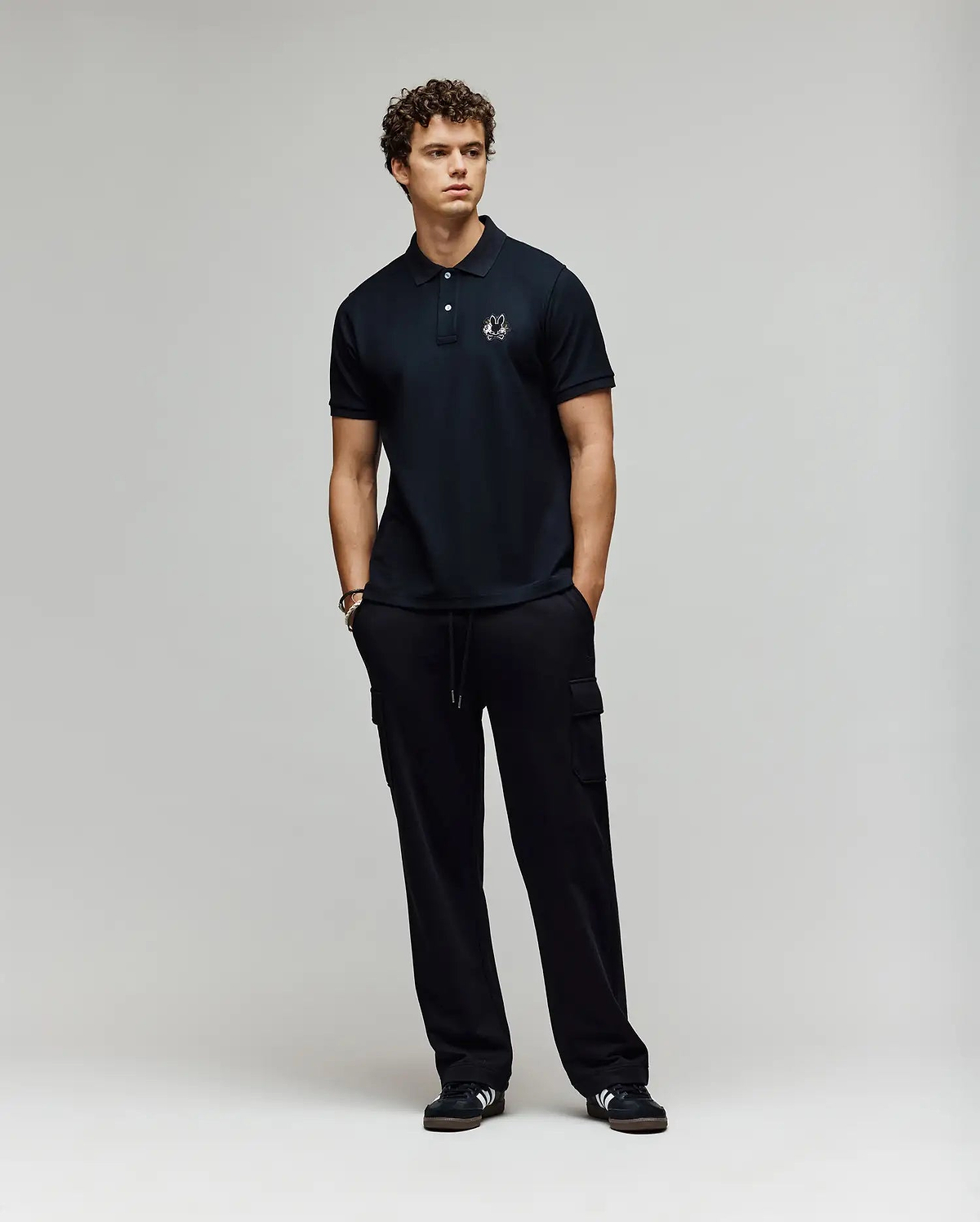 A young man with curly hair stands against a plain background, wearing a Psycho Bunny MENS AUGUSTE PIQUE POLO SHIRT - MT0100051 and black cargo pants. Black sneakers with white accents complete his varsity-inspired look as he gazes to the side. (4)