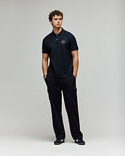 A young man with curly hair stands against a plain background, wearing a Psycho Bunny MENS AUGUSTE PIQUE POLO SHIRT - MT0100051 and black cargo pants. Black sneakers with white accents complete his varsity-inspired look as he gazes to the side. (4)