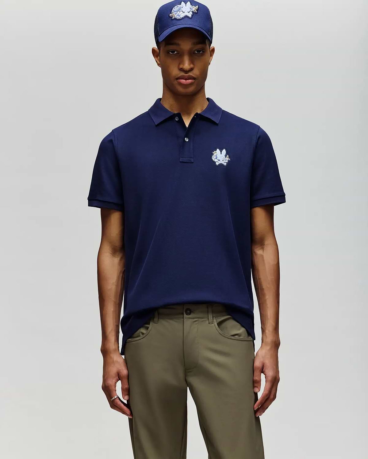 A man in Psycho Bunny’s MENS AUGUSTE PIQUE POLO SHIRT (MT0100051), navy blue, stands against a light background, paired with khaki pants and a matching navy cap. (3)