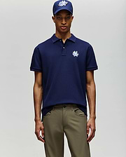 A man in Psycho Bunny’s MENS AUGUSTE PIQUE POLO SHIRT (MT0100051), navy blue, stands against a light background, paired with khaki pants and a matching navy cap. (3)