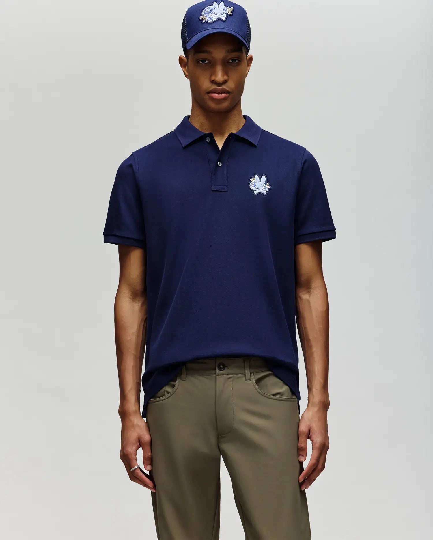 A man in Psycho Bunny’s MENS AUGUSTE PIQUE POLO SHIRT (MT0100051), navy blue, stands against a light background, paired with khaki pants and a matching navy cap. (3)