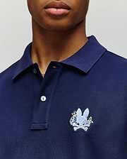 A person wearing the Psycho Bunny MENS AUGUSTE PIQUE POLO SHIRT (MT0100051) in navy blue, featuring a collared neckline, button placket, and a large embroidered rabbit head over crossed bones on the left chest for varsity flair. (2)
