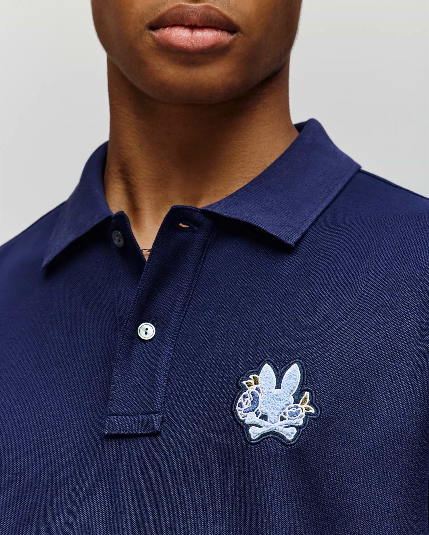 A person wearing the Psycho Bunny MENS AUGUSTE PIQUE POLO SHIRT (MT0100051) in navy blue, featuring a collared neckline, button placket, and a large embroidered rabbit head over crossed bones on the left chest for varsity flair. (2)