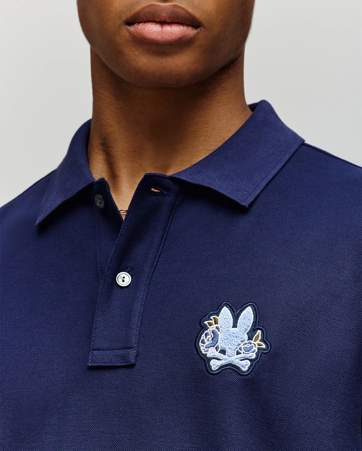 A person wearing the Psycho Bunny MENS AUGUSTE PIQUE POLO SHIRT (MT0100051) in navy blue, featuring a collared neckline, button placket, and a large embroidered rabbit head over crossed bones on the left chest for varsity flair. (2)