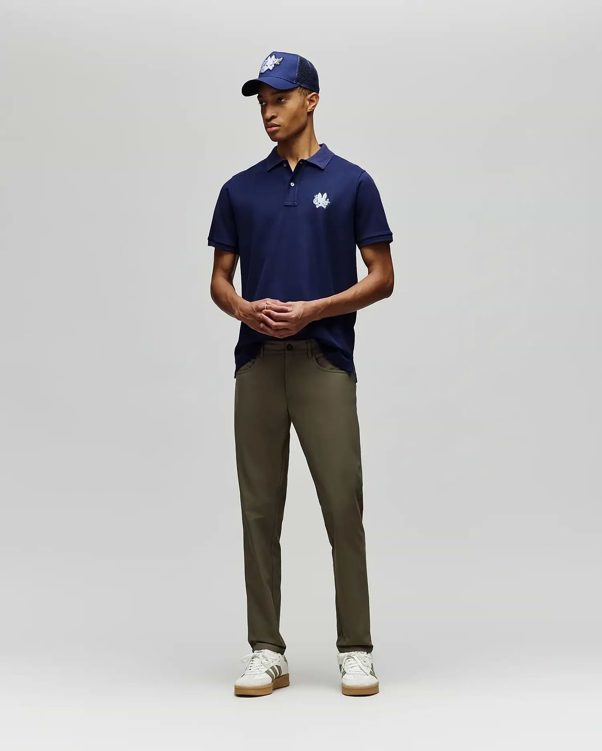 A man stands against a plain background wearing the Psycho Bunny MENS AUGUSTE PIQUE POLO SHIRT - MT0100051 in navy with a white logo, a matching cap, olive green pants, and white sneakers. His hands are loosely clasped in front of him. (4)