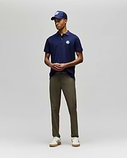 A man stands against a plain background wearing the Psycho Bunny MENS AUGUSTE PIQUE POLO SHIRT - MT0100051 in navy with a white logo, a matching cap, olive green pants, and white sneakers. His hands are loosely clasped in front of him. (4)
