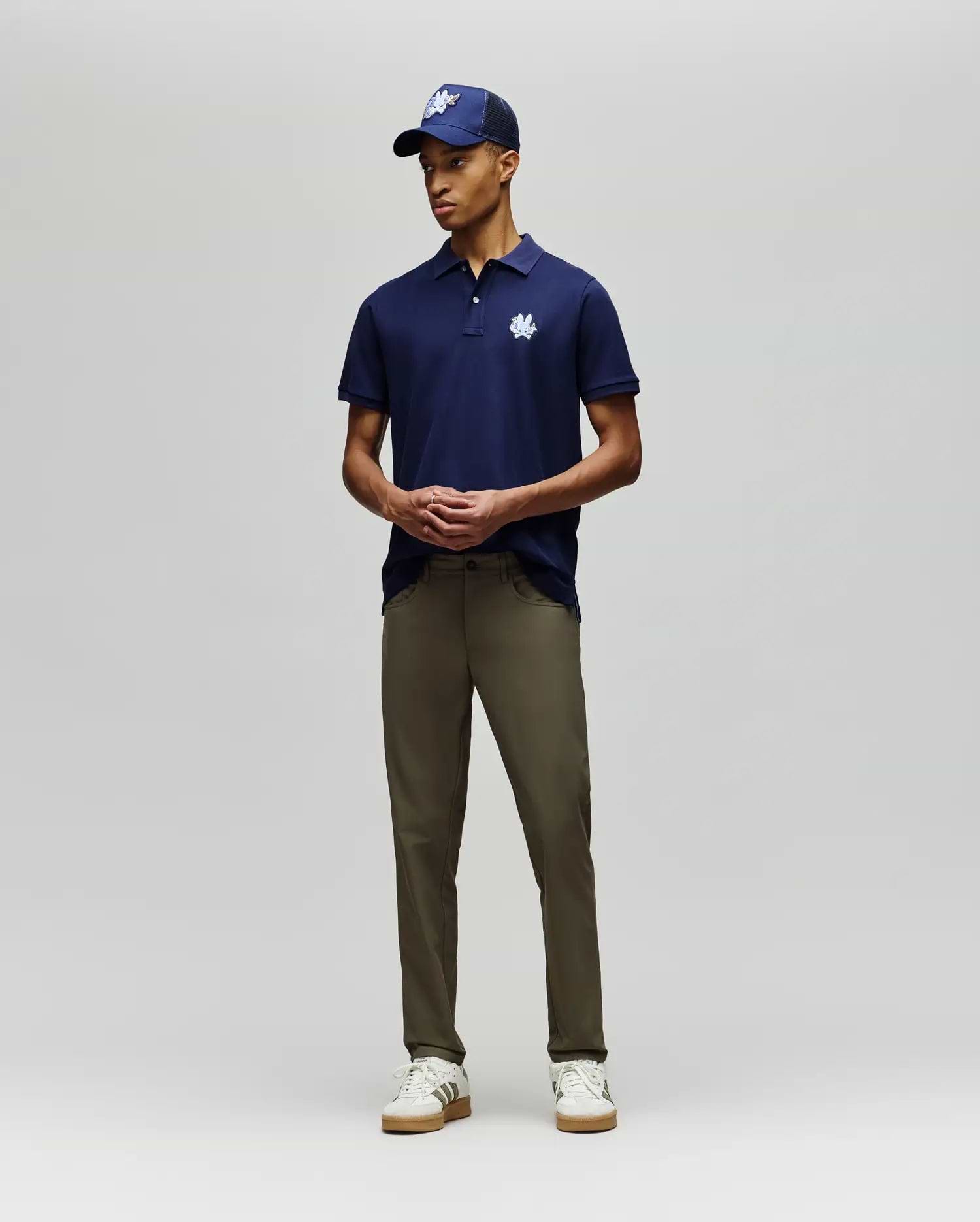 A man stands against a plain background wearing the Psycho Bunny MENS AUGUSTE PIQUE POLO SHIRT - MT0100051 in navy with a white logo, a matching cap, olive green pants, and white sneakers. His hands are loosely clasped in front of him. (4)