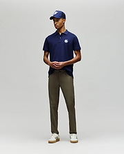 A man stands against a plain background wearing the Psycho Bunny MENS AUGUSTE PIQUE POLO SHIRT - MT0100051 in navy with a white logo, a matching cap, olive green pants, and white sneakers. His hands are loosely clasped in front of him. (4)