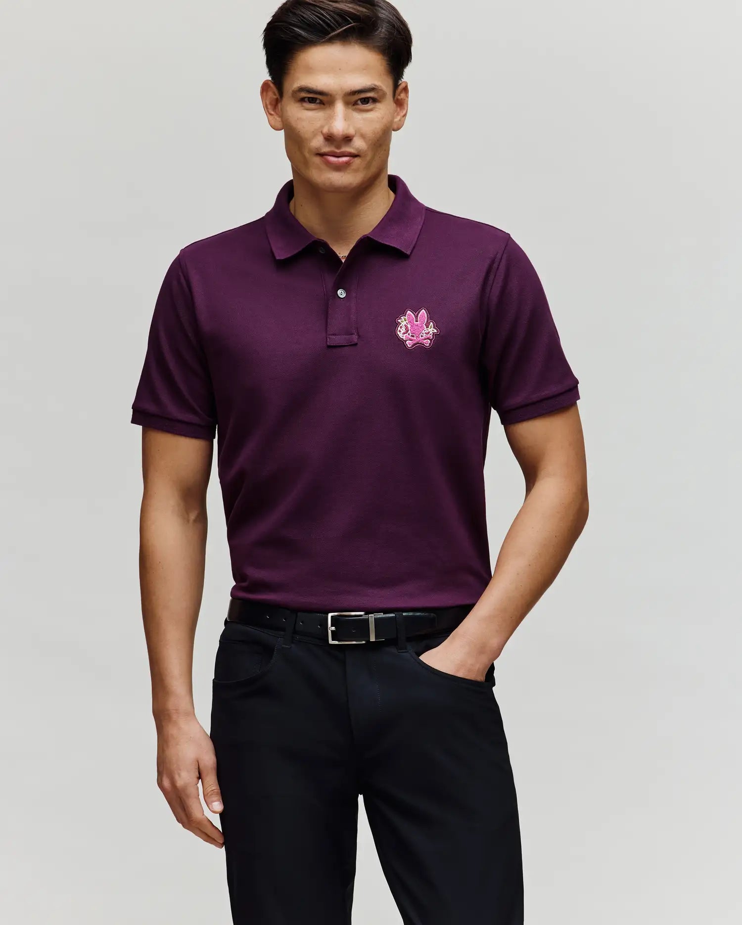 A man stands against a plain background wearing the Psycho Bunny MENS AUGUSTE PIQUE POLO SHIRT - MT0100051, a short-sleeved, dark purple Pima cotton polo with a small chest logo, paired with black pants and a black belt.