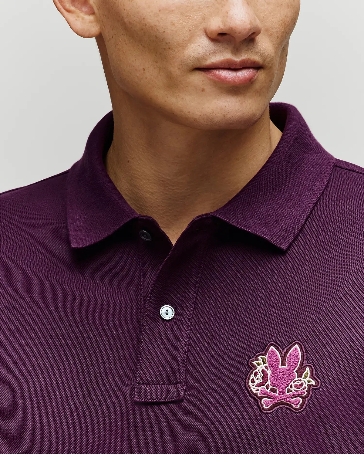 A person wears the Psycho Bunny MENS AUGUSTE PIQUE POLO SHIRT (MT0100051) in dark purple, featuring a collar and a chest patch with the brand's iconic pink bunny and crossed bones logo. The photo highlights this standout Americana style piece.