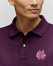 A person wears the Psycho Bunny MENS AUGUSTE PIQUE POLO SHIRT (MT0100051) in dark purple, featuring a collar and a chest patch with the brand's iconic pink bunny and crossed bones logo. The photo highlights this standout Americana style piece.