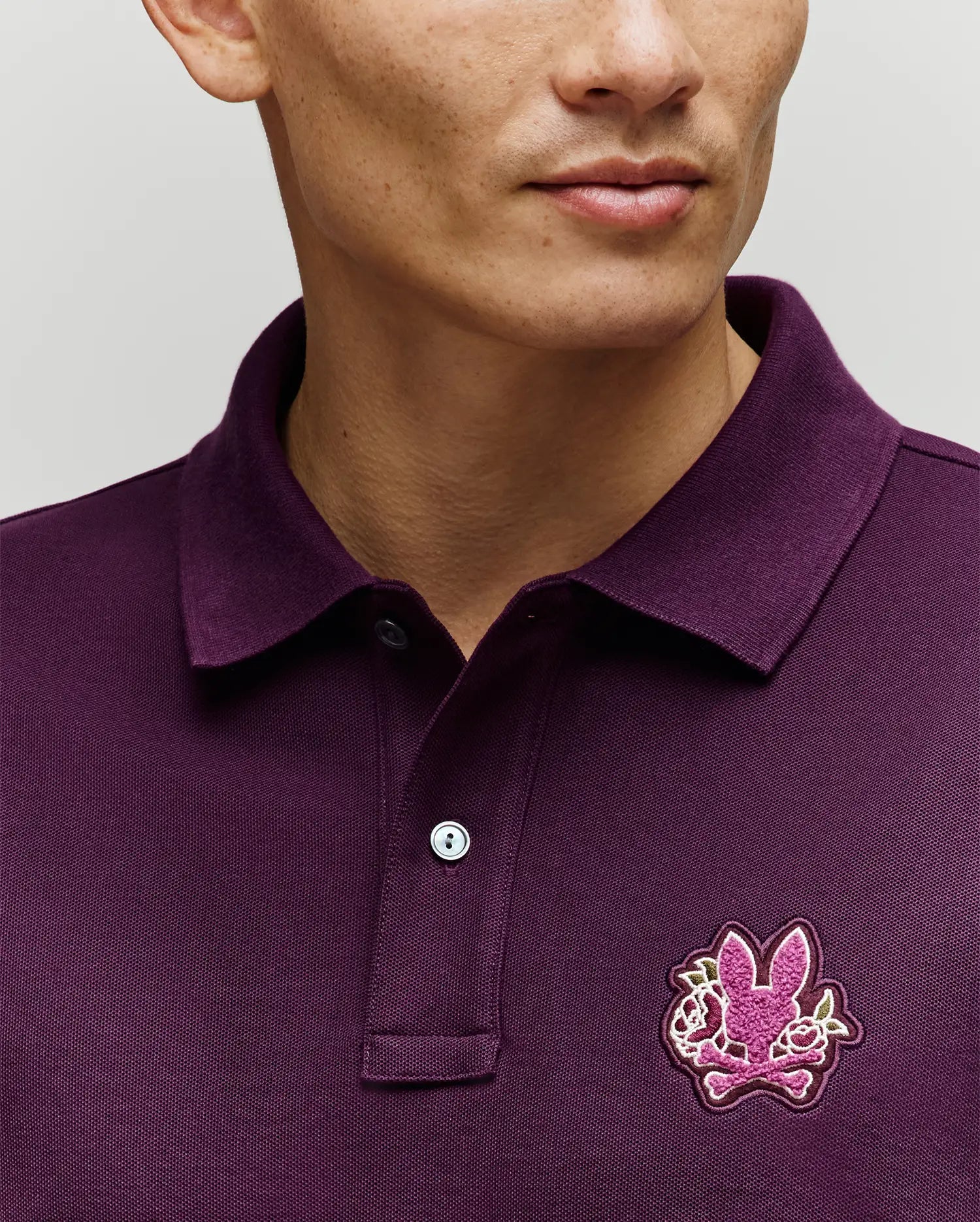A person wears the Psycho Bunny MENS AUGUSTE PIQUE POLO SHIRT (MT0100051) in dark purple, featuring a collar and a chest patch with the brand's iconic pink bunny and crossed bones logo. The photo highlights this standout Americana style piece.