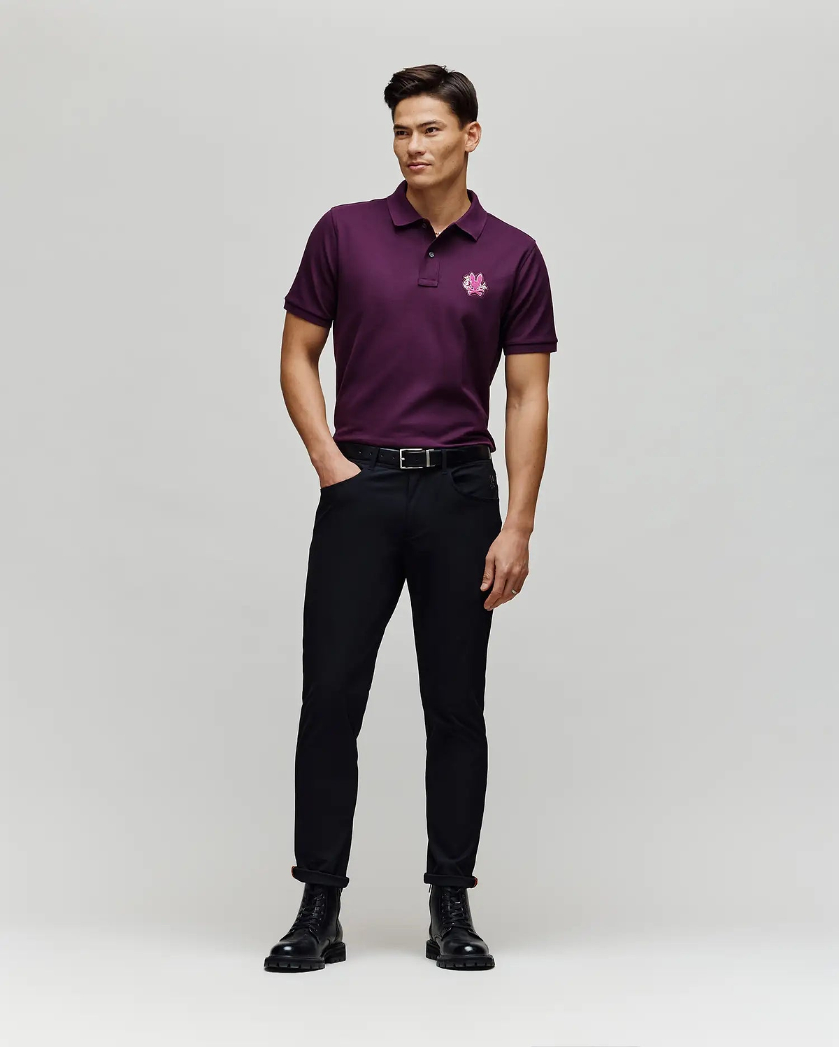 A man stands in a studio wearing the Psycho Bunny MENS AUGUSTE PIQUE POLO SHIRT - MT0100051 in dark purple, paired with black pants and boots. He smiles, looking to the side with one hand in his pocket against a plain light gray background.