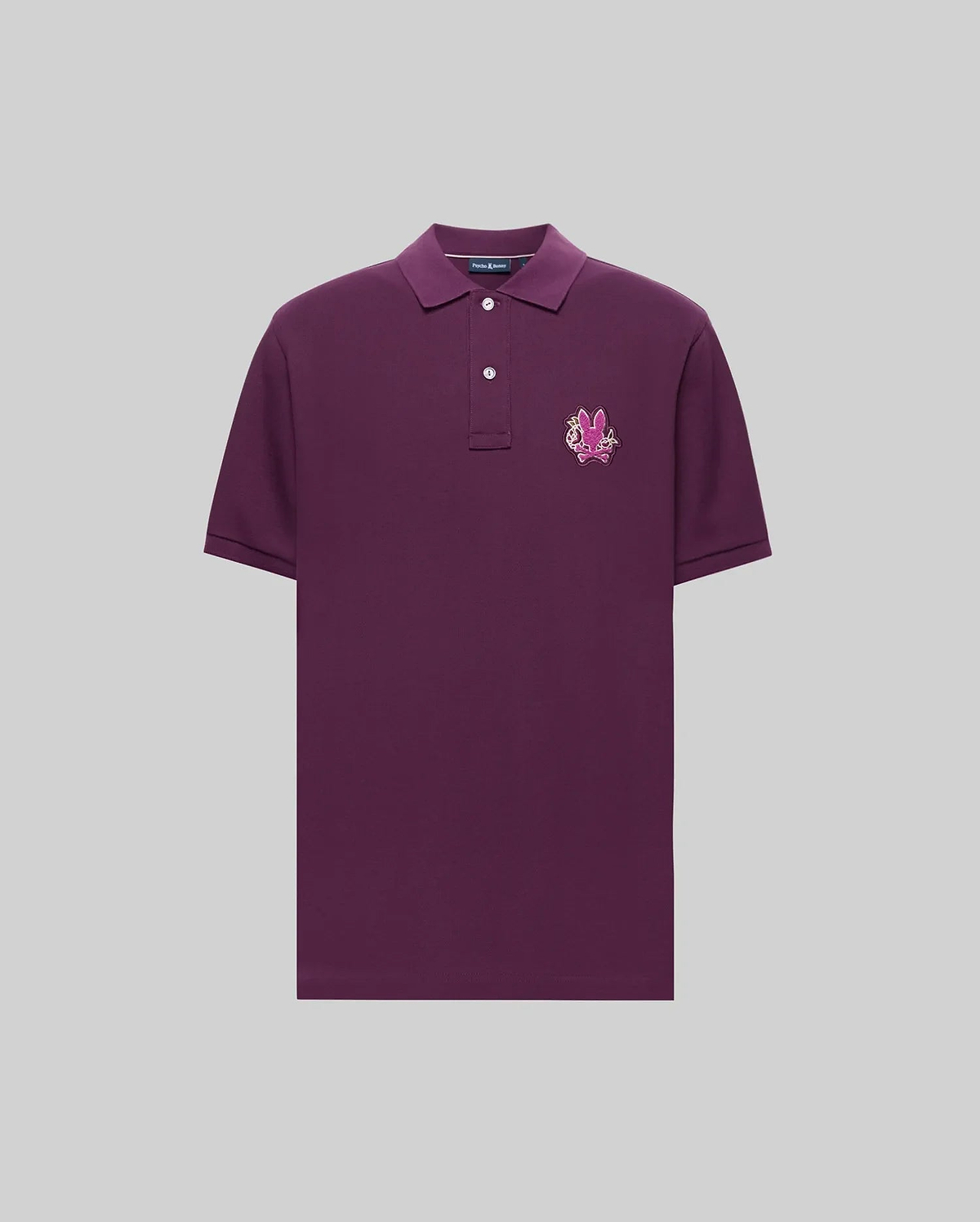 The Psycho Bunny MENS AUGUSTE PIQUE POLO SHIRT (MT0100051) is a short-sleeved, dark purple Pima cotton polo with a two-button collar and a pink-white embroidered crest on the left chest, shown against a light gray background.